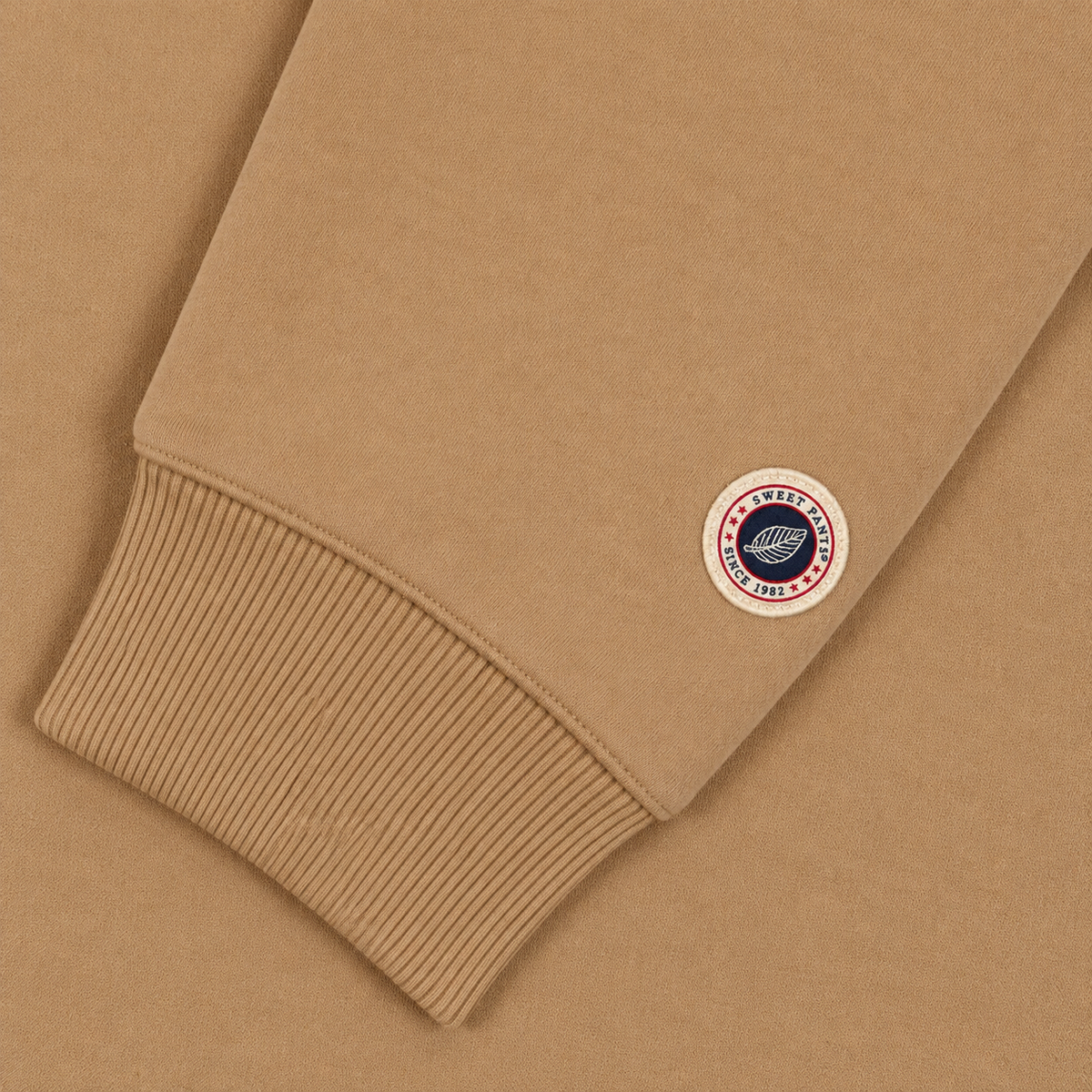 Gros plan sur le patch logo tricolore Sweet Pants au bas de la manche du sweat beige et finition bord-côte