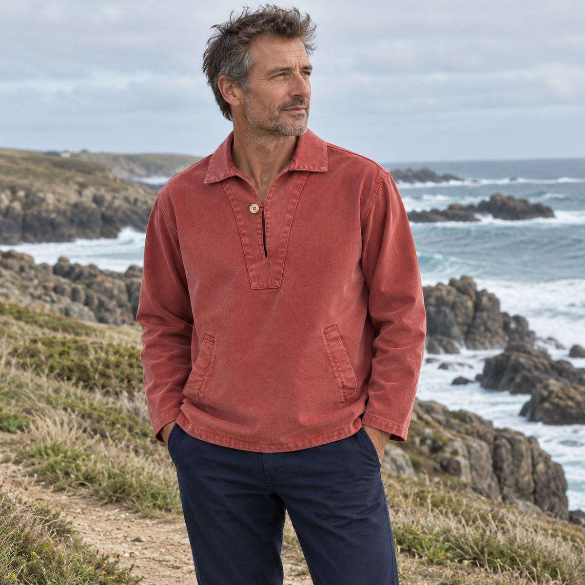 Homme portant la vareuse DINARD RUST PULLIN rouge brique avec un pantalon marine, sur un sentier côtier face à l’océan.